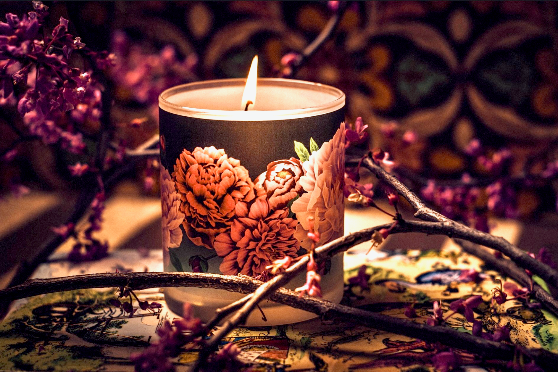Peonies Candle with bold floral design in reds and pinks lit against a black background.