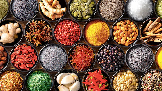 Overhead view of assorted spices and ingredients in small black bowls, including turmeric, ginger, star anise, peppercorns, seeds, dried chilies, garlic, and coarse salts arranged on a wooden surface.